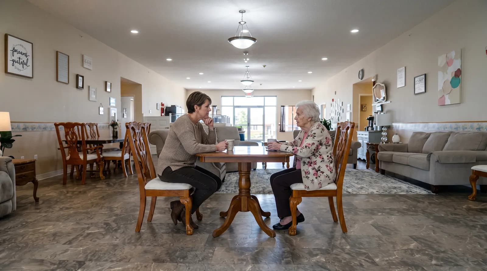 Illustration: an adult daughter and her mother sitting across a small table, talking, in the open living and dining room of a small assisted living home.