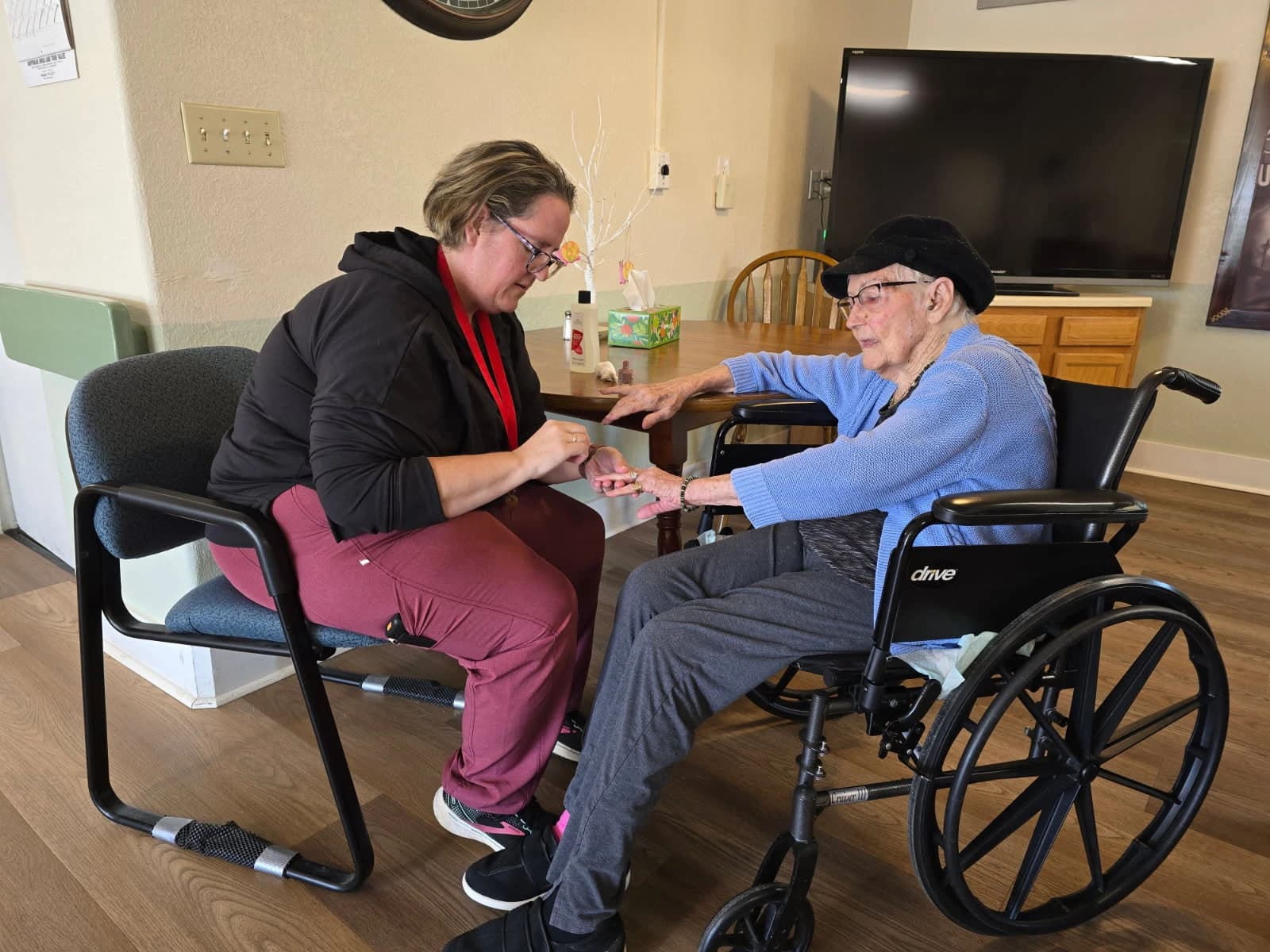 An Aspen Leaf caregiver sitting with an elderly resident in a wheelchair, helping her in a warm, home-like common room